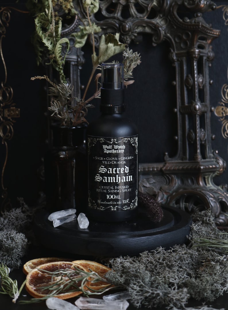 May include: A black glass bottle with a spray nozzle, labeled "Wolf Wythch Apothecary" and "Sacred Samhain Crystal Infused Ritual Saining Spray 100ML Handmade in the UK". The bottle is surrounded by dried orange slices, clear quartz crystals, and green moss.