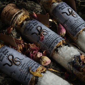Op de afbeelding: Drie glazen flesjes gevuld met badzout en gedroogde bloemen. De flesjes zijn gelabeld met "Mama Moon Bath Shot" en hebben een slangendesign. De labels bevatten ook de tekst "www.wolfwitch.com".