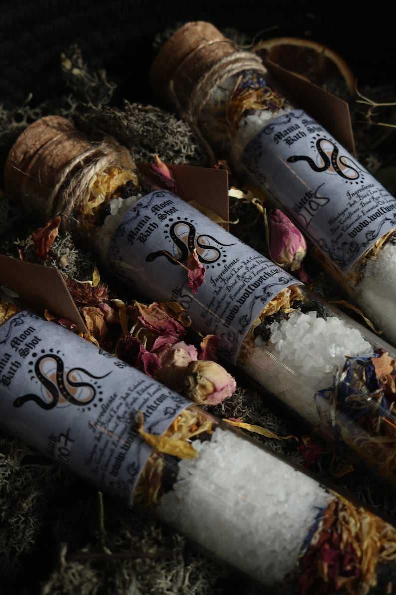 Op de afbeelding: Drie glazen flesjes gevuld met badzout en gedroogde bloemen. De flesjes zijn gelabeld met "Mama Moon Bath Shot" en hebben een slangendesign. De labels bevatten ook de tekst "www.wolfwitch.com".