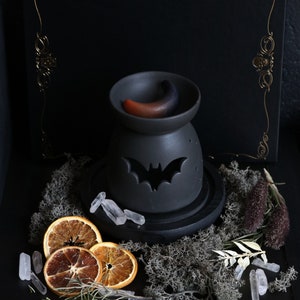 May include: Black ceramic oil burner with a bat design and a crescent moon shaped wax melt. The burner is on a black wooden base and surrounded by dried orange slices, crystals, and dried moss.