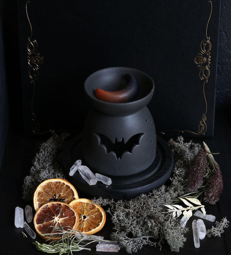 May include: Black ceramic oil burner with a bat design and a crescent moon shaped wax melt. The burner is on a black wooden base and surrounded by dried orange slices, crystals, and dried moss.