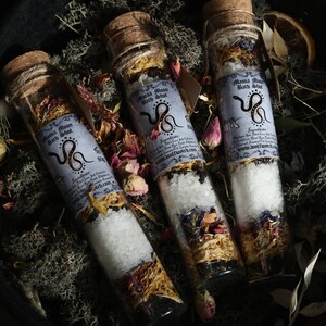 Op de afbeelding: Drie glazen flesjes gevuld met badzout en gedroogde bloemen. De flesjes hebben kurken stoppen en etiketten met de tekst "Mama Moon Bath Shot" met een slangillustratie. De flesjes zijn gerangschikt op een bed van mos en gedroogde bloemen.