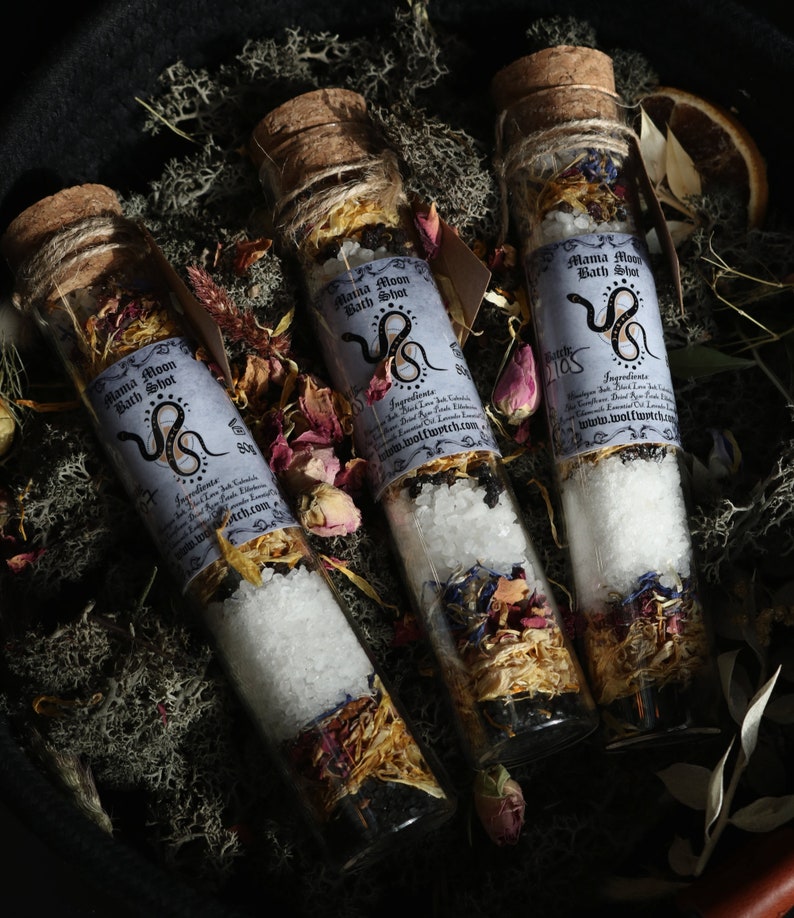 Op de afbeelding: Drie glazen flesjes gevuld met badzout en gedroogde bloemen. De flesjes hebben kurken stoppen en etiketten met de tekst "Mama Moon Bath Shot" met een slangillustratie. De flesjes zijn gerangschikt op een bed van mos en gedroogde bloemen.