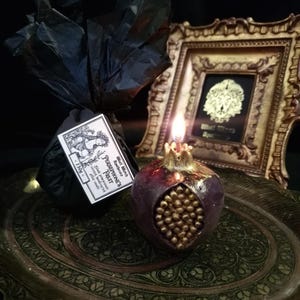 May include: A lit pomegranate-shaped candle with gold accents sits on a decorative tray. The candle is labelled "Persephone's Fruit" and "Black Witch Apothecary".