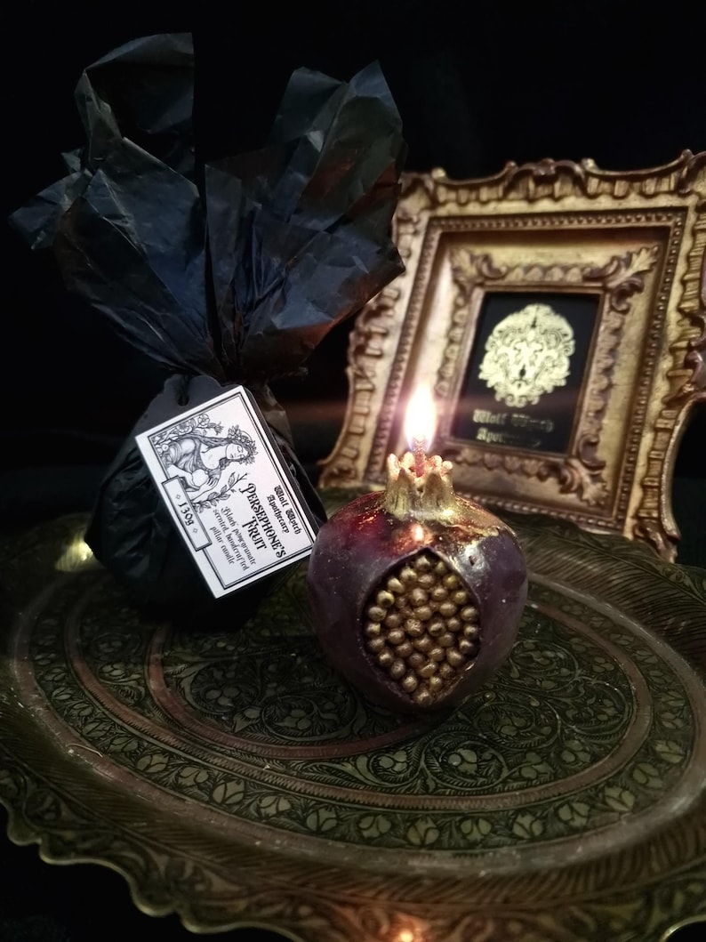 May include: A lit pomegranate-shaped candle with gold accents sits on a decorative tray. The candle is labelled "Persephone's Fruit" and "Black Witch Apothecary".