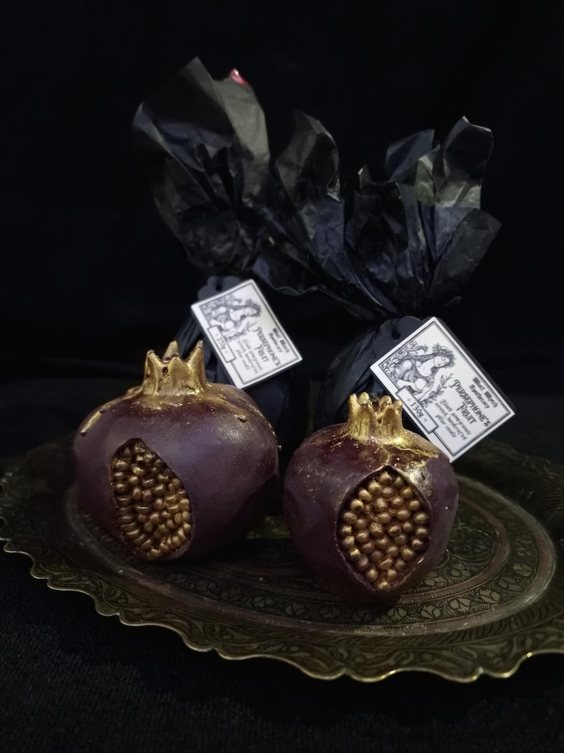 May include: Two dark purple pomegranate-shaped candles with gold detailing on a gold tray. The candles are wrapped in black tissue paper with a label that reads "Pomegranate Fruit" in a decorative font.