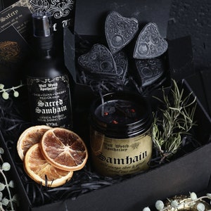 May include: A black gift box filled with a black candle, a bottle of spray, dried orange slices, and black heart-shaped charms. The candle label reads "Wolf Wytch Apothecary Samhain Wild orange clove & ginger Coconut wax candle". The spray bottle label reads "Wolf Wytch Apothecary Sacred Samhain Crystal Infused Ritual Saining Spray 100ml".
