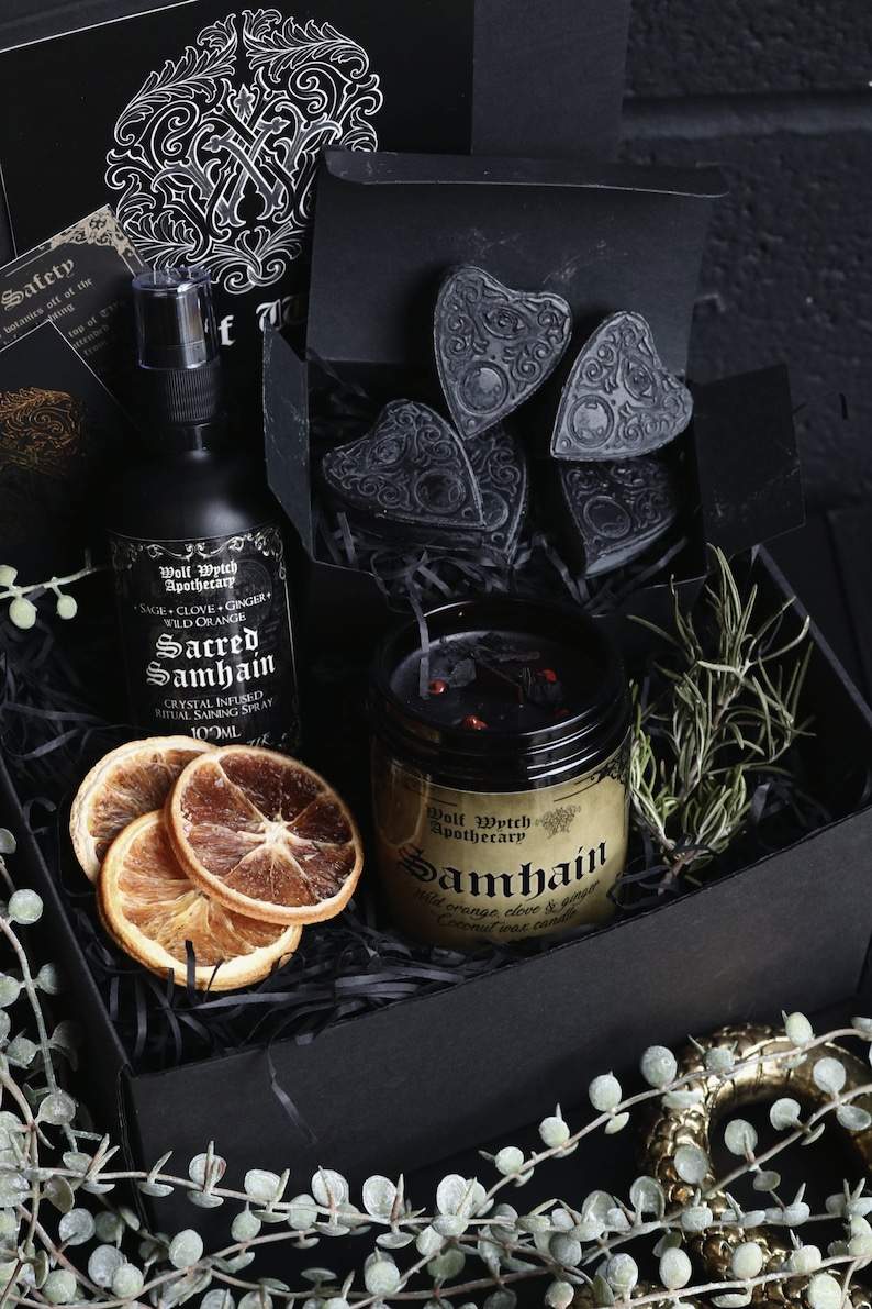May include: A black gift box filled with a black candle, a bottle of spray, dried orange slices, and black heart-shaped charms. The candle label reads "Wolf Wytch Apothecary Samhain Wild orange clove & ginger Coconut wax candle". The spray bottle label reads "Wolf Wytch Apothecary Sacred Samhain Crystal Infused Ritual Saining Spray 100ml".