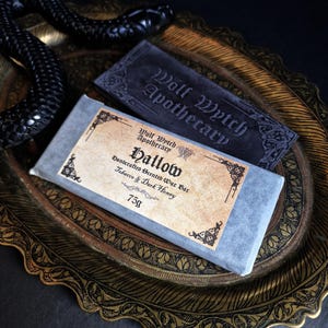 May include: A handcrafted scented wax bar labeled "Hallow" with "Tobacco & Dark Honey" scent, and a dark gray soap bar from "Wolf Wytch Apothecary". The bars rest on a decorative gold-toned tray with a black snake.