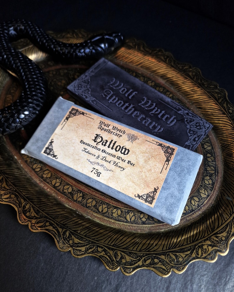 May include: A handcrafted scented wax bar labeled "Hallow" with "Tobacco & Dark Honey" scent, and a dark gray soap bar from "Wolf Wytch Apothecary". The bars rest on a decorative gold-toned tray with a black snake.