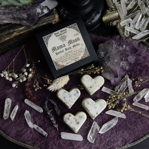 Op de afbeelding: Vier hartvormige badmelts in wit en roze met gedroogde bloemen en kruiden. De melts liggen op een paarse ondergrond met heldere kwartskristallen en een zwarte doos met de tekst "Mama Moon Herbal Bath Melts" en een heksenketel-logo.
