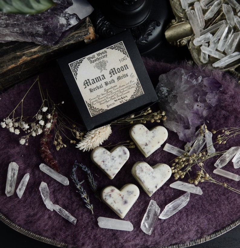 Op de afbeelding: Vier hartvormige badmelts in wit en roze met gedroogde bloemen en kruiden. De melts liggen op een paarse ondergrond met heldere kwartskristallen en een zwarte doos met de tekst "Mama Moon Herbal Bath Melts" en een heksenketel-logo.