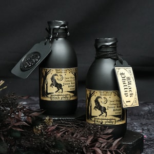 Può includere: Due bottiglie di vetro nero con gocce di cera nera sulla parte superiore. Le bottiglie hanno etichette nere e dorate con un'illustrazione di capra e il testo "Wolf Wytch Apothecary" e "Black Phillip". Le bottiglie sono appoggiate su una superficie nera con fiori secchi.