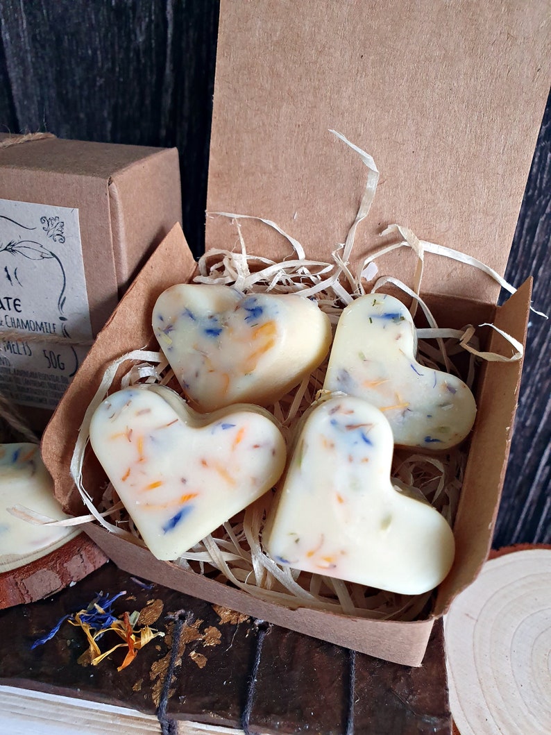 May include: Heart-shaped wax melts with colorful floral inclusions are displayed in a brown cardboard box with shredded paper. The melts are creamy white and the box is open, revealing the contents. A second box is partially visible.