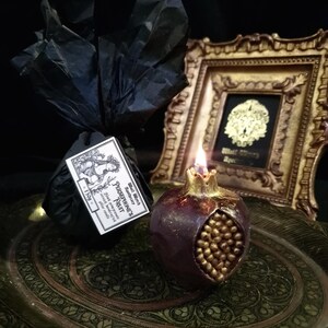 May include: A dark purple pomegranate-shaped candle with gold details sits on a decorative tray. The candle is lit and has a small flame. The candle is labelled "Persephone's Fruit" with the text "Small Batch Soy Wax" and "Black Raspberry & Plum Scented Candle".