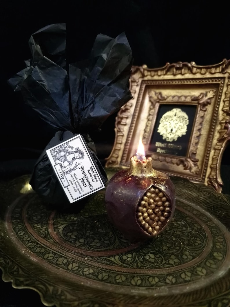 May include: A dark purple pomegranate-shaped candle with gold details sits on a decorative tray. The candle is lit and has a small flame. The candle is labelled "Persephone's Fruit" with the text "Small Batch Soy Wax" and "Black Raspberry & Plum Scented Candle".