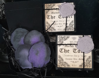 May include: Two boxes of Morbid Apothecary coconut wax melts, each with a purple wax seal. The boxes are labelled "The Tower" and feature a black and white design with the text "Blueberries, Rose & Vanilla". The wax melts are in a black box with black shredded paper.