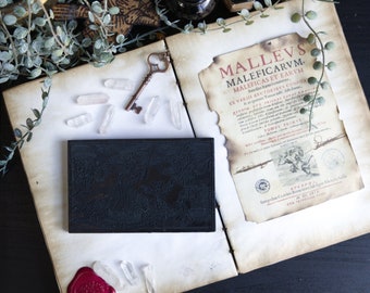 May include: An open book with a page titled "Mallevs Maleficarum et Earum" with a black and white illustration of a scene from the book. The book is surrounded by crystals, a key, and a wax seal.