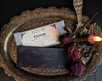 May include: A still-life image featuring two dark gray soap bars and a decorative metal tray. One soap bar is wrapped in paper with the text "Hallow" and "Tobacco & Dark Honey". The other bar is embossed with "Wolf Wytch Apothecary".