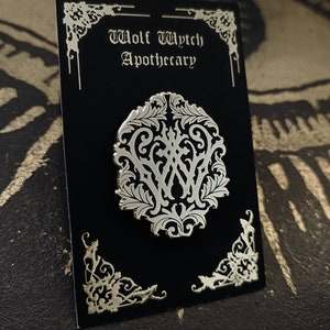 May include: A silver-colored pin with a decorative design featuring the letters "WWA" surrounded by intricate floral patterns. The pin is displayed on a black card with the text "Wolf Wytch Apothecary" in a gothic font.