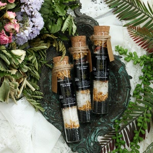 May include: Three glass test tubes filled with bath salts and dried flowers. The tubes are labeled "Winter Solstice Bath Salt". The tubes are on a green and brown metal tray.