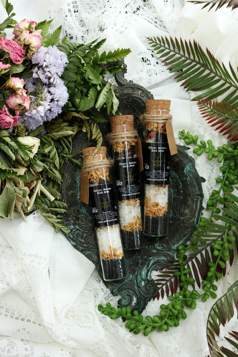 May include: Three glass test tubes filled with bath salts and dried flowers. The tubes are labeled "Winter Solstice Bath Salt". The tubes are on a green and brown metal tray.