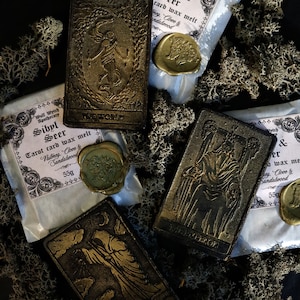May include: Four tarot card wax melts with gold foil accents and a black background. The cards are labeled "The World", "The Hermit", "Sibyl & Seer", and "Sibyl & Seer". Each card has a wax seal with a gold design.