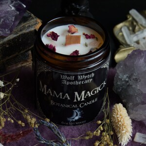 May include: A brown glass candle jar with a black label that reads "Wolf Wytch Apothecary Mama Magick Botanical Candle 6oz Net Weight". The candle is white and has dried rose petals and a wooden wick.