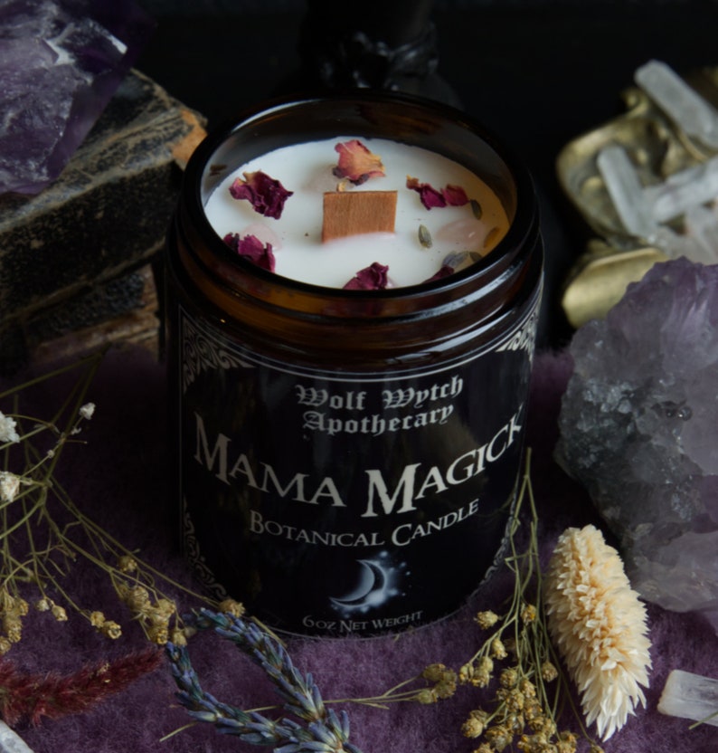 May include: A brown glass candle jar with a black label that reads "Wolf Wytch Apothecary Mama Magick Botanical Candle 6oz Net Weight". The candle is white and has dried rose petals and a wooden wick.