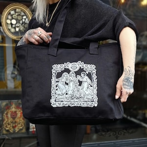 May include: Black tote bag with a white illustration of three women dancing within a tree branch frame. The bag has two black handles and is made of a durable fabric. The design is centered on the front of the bag.