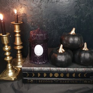 May include: Three black pumpkins with gold stems sit on top of a stack of three books. The books have a black and gold design with the words "Beyond the Dark Veil" on the bottom book. Two gold candle holders with black candles are on the left side of the image. A black candle with a white wax seal is in front of the books.
