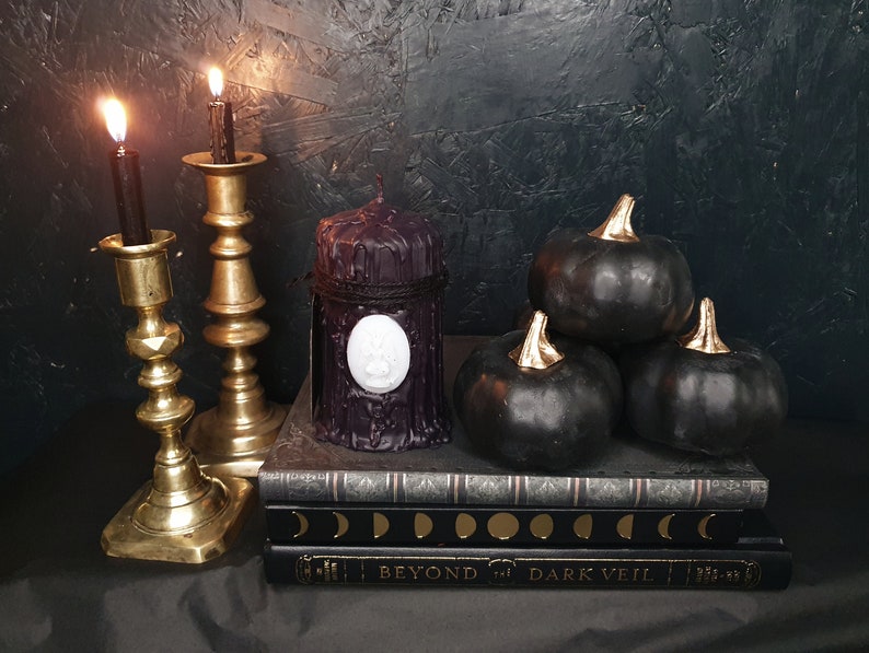 May include: Three black pumpkins with gold stems sit on top of a stack of three books. The books have a black and gold design with the words "Beyond the Dark Veil" on the bottom book. Two gold candle holders with black candles are on the left side of the image. A black candle with a white wax seal is in front of the books.