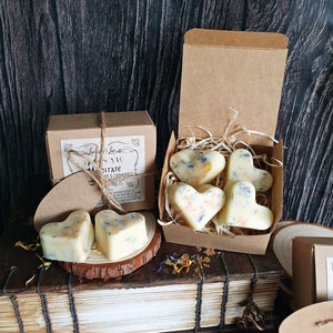 May include: Heart-shaped wax melts with blue and yellow floral accents are displayed in a brown cardboard box and on a wooden surface. The box is labeled "Meditate" with the brand name "Luminare Lux". The background is a dark wood.