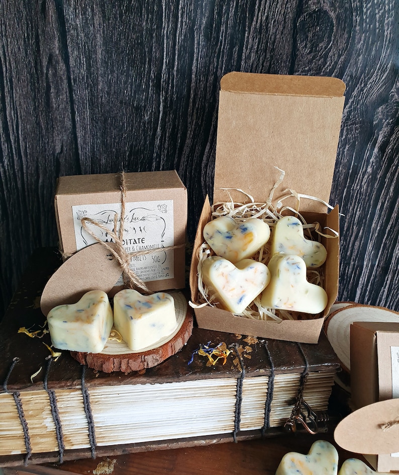 May include: Heart-shaped wax melts with blue and yellow floral accents are displayed in a brown cardboard box and on a wooden surface. The box is labeled "Meditate" with the brand name "Luminare Lux". The background is a dark wood.