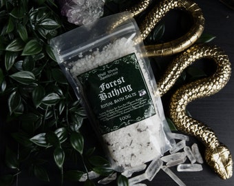 以下が含まれることがあります： 白いバスソルトが入った透明なプラスチック袋。袋には「Wolf Witch Apothecary Forest Bathing Ritual Bath Salts 300g」と書かれた緑色のラベルが付いています。袋は緑の葉、透明な水晶、金色の蛇の彫刻で囲まれています。