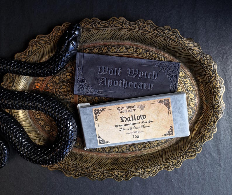 May include: A dark, ornate tray holds two bars of soap. One is a dark gray rectangle with "Wolf Wytch Apothecary" embossed on it. The other is a rectangular bar of soap in a gray box with a label that reads "Hallow" and "75g". A black snake decorates the tray.