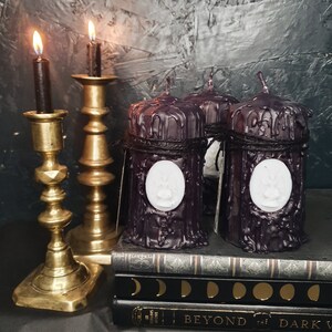 May include: Four black pillar candles with a white wax design on each. The candles are on a stack of books with the words "Beyond the Dark Veil" on the front.