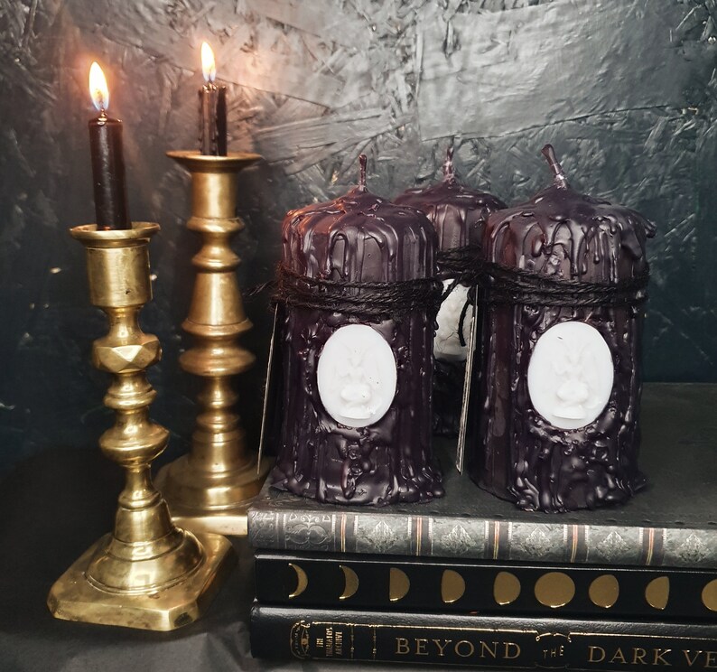 May include: Four black pillar candles with a white wax design on each. The candles are on a stack of books with the words "Beyond the Dark Veil" on the front.