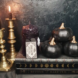 May include: Three black pumpkins with gold stems are arranged on top of a stack of books. The books have a black cover with gold lettering that reads "Beyond the Dark Veil". A black candle with a white label that reads "Hallow" sits on the books. Two gold candle holders with lit candles are in the background.