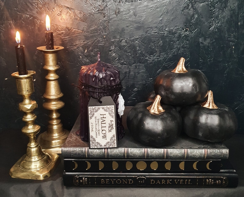 May include: Three black pumpkins with gold stems are arranged on top of a stack of books. The books have a black cover with gold lettering that reads "Beyond the Dark Veil". A black candle with a white label that reads "Hallow" sits on the books. Two gold candle holders with lit candles are in the background.