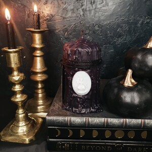 May include: Black pillar candle with a white wax paw print design, sitting on a stack of books with a dark background. The candle is surrounded by black decorative gourds.
