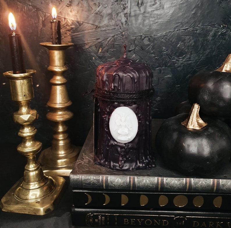 May include: Black pillar candle with a white wax paw print design, sitting on a stack of books with a dark background. The candle is surrounded by black decorative gourds.