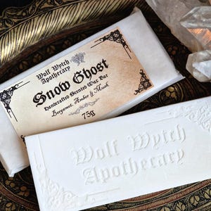 K&ouml;nnte beinhalten: Zwei wei&szlig;e Wachsriegel mit dem Text "Wolf Wytch Apothecary" und "Snow Ghost" auf der Verpackung. Die Riegel liegen auf einem dekorativen, goldfarbenen Tablett. Der "Snow Ghost" Riegel wiegt 75g und ist mit Bergamotte, Amber & Moschus parf&uuml;miert.