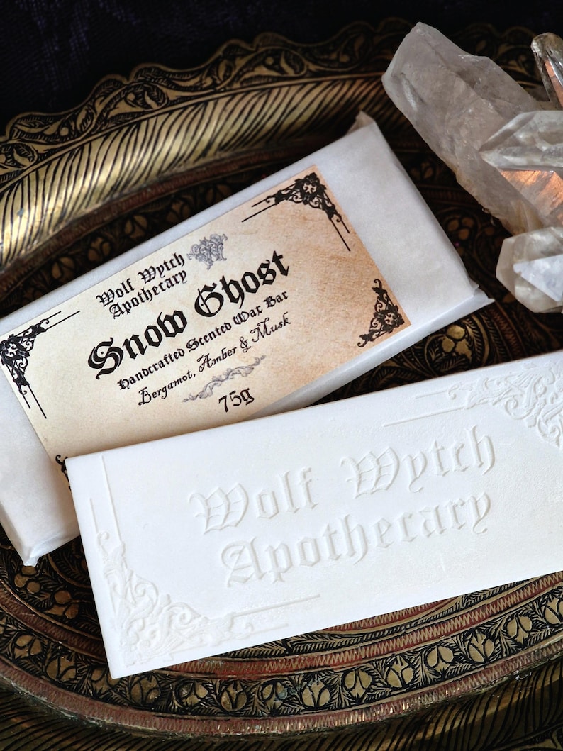 K&ouml;nnte beinhalten: Zwei wei&szlig;e Wachsriegel mit dem Text "Wolf Wytch Apothecary" und "Snow Ghost" auf der Verpackung. Die Riegel liegen auf einem dekorativen, goldfarbenen Tablett. Der "Snow Ghost" Riegel wiegt 75g und ist mit Bergamotte, Amber & Moschus parf&uuml;miert.