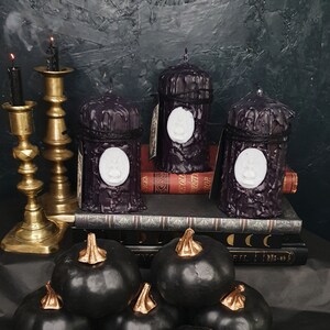 May include: Three black pillar candles with white raised designs on a stack of books. The candles are on a black surface with a gold metal candle holder and black pumpkins in the foreground.