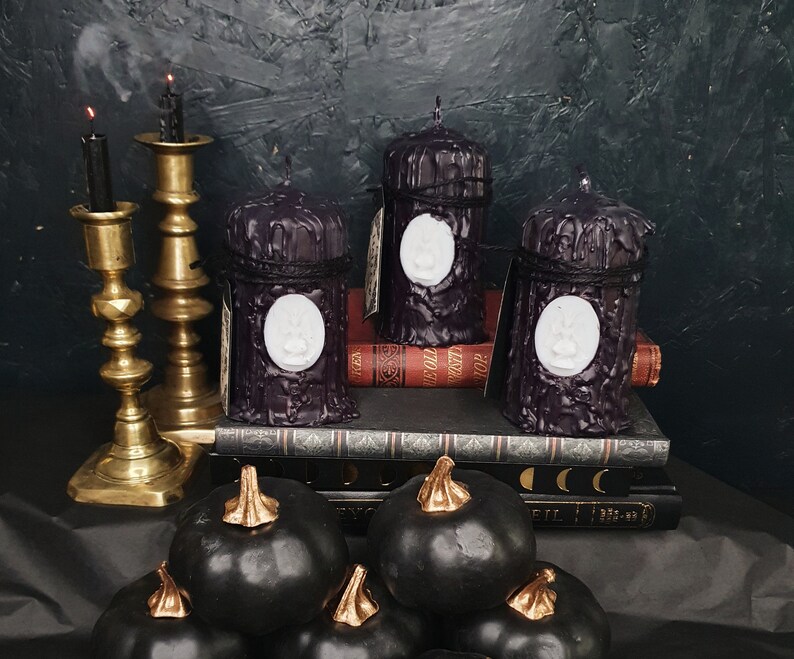 May include: Three black pillar candles with white raised designs on a stack of books. The candles are on a black surface with a gold metal candle holder and black pumpkins in the foreground.