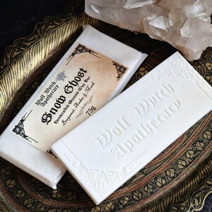 K&ouml;nnte beinhalten: Zwei wei&szlig;e Wachsriegel, einer mit einem Etikett mit der Aufschrift "Snow Ghost" und "Wolf Wytch Apothecary" verpackt. Der unverpackte Riegel ist mit "Wolf Wytch Apothecary" gepr&auml;gt. Die Riegel liegen auf einem dekorativen, verzierten Tablett mit einem Kristallcluster im Hintergrund.