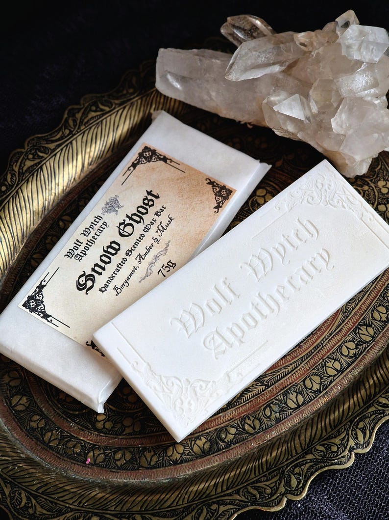 K&ouml;nnte beinhalten: Zwei wei&szlig;e Wachsriegel, einer mit einem Etikett mit der Aufschrift "Snow Ghost" und "Wolf Wytch Apothecary" verpackt. Der unverpackte Riegel ist mit "Wolf Wytch Apothecary" gepr&auml;gt. Die Riegel liegen auf einem dekorativen, verzierten Tablett mit einem Kristallcluster im Hintergrund.