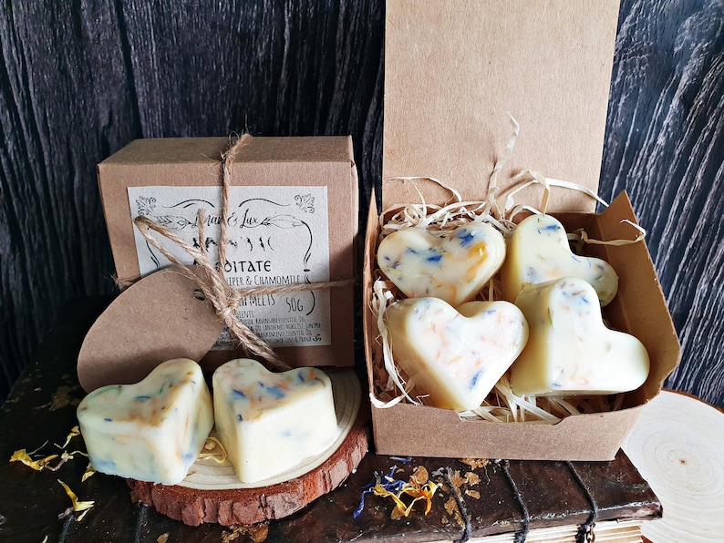 May include: Heart-shaped wax melts with blue and orange floral accents are displayed in a brown box and on a wooden slice. The box is labeled "Meditate" with Juniper & Chamomile. The melts are a creamy white color.
