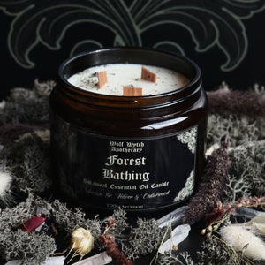 K&ouml;nnte beinhalten: Eine dunkelbraune Glasgef&auml;&szlig;kerze mit einem schwarzen Etikett, auf dem "Wolf Wytch Apothecary Forest Bathing Botanical Essential Oil Candle Siberian Fir, Vetiver & Cedarwood 500g NET WEIGHT" steht. Die Kerze hat einen wei&szlig;en Wachsoberteil mit drei Holzdochten.